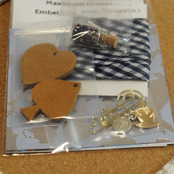 Back of Alice in Wonderland Mini Kit with bead vial, plaid ribbon, leather heart charms, and gold clock and hat charms visible through clear packaging.