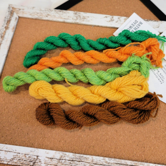 Five skeins of Bathhouse Dyeworks  tapestry yarn in green, orange, light green, yellow, brown, and dark brown, arranged on a cork board inside a distressed white wooden frame. 