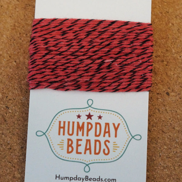 Red and black hand dyed bakers twine on a Humpday Beads label against a cork background