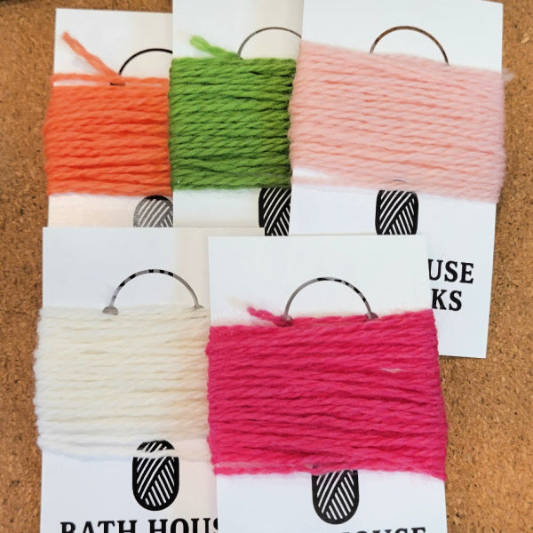 Five skeins of yarn in orange, green, pale pink, and cream, each mounted on a white card, arranged on a brown cork-like background. 