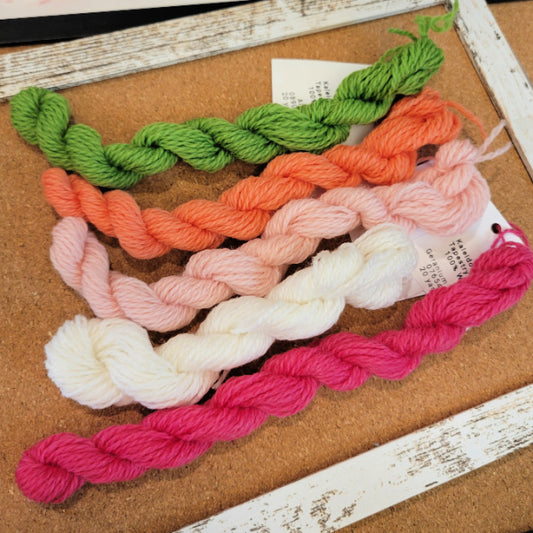Five tapestry yarn skeins in green, orange, light pink, white, and bright pink, arranged diagonally on a cork board with a rustic white wooden frame.